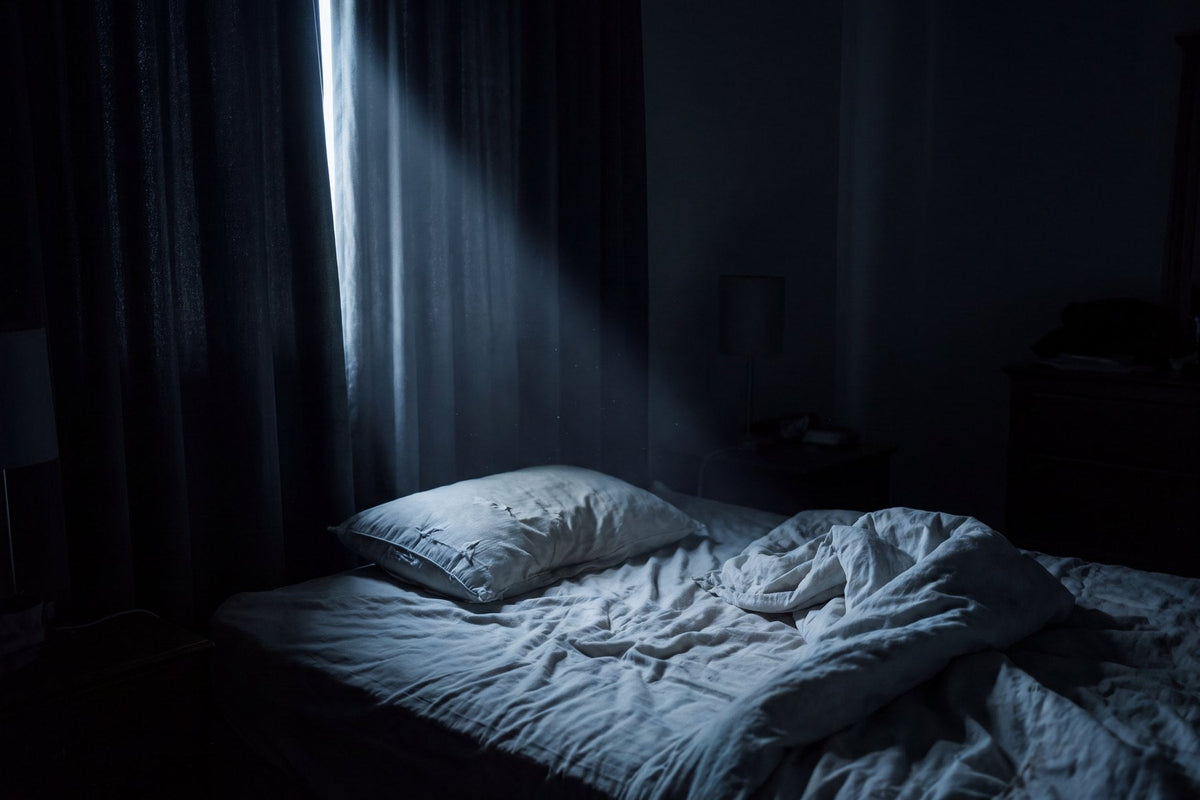 An unmade bed in a dark bedroom lit only by a thin sliver of moonlight through the curtains