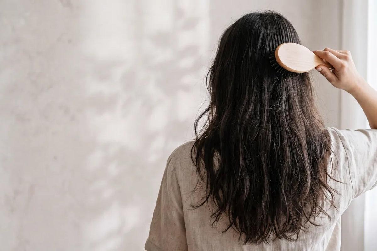 A woman photographed from behind, brushing her long dark hair in soft morning window light