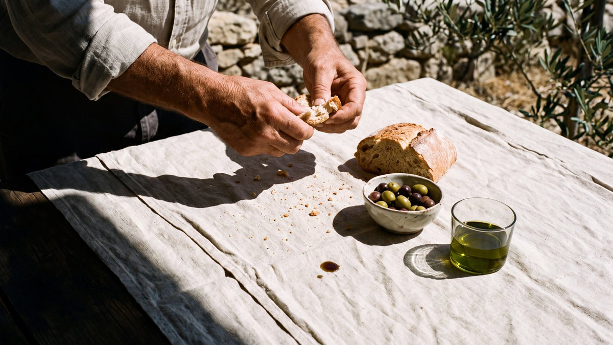 Hands tearing fresh bread at a Mediterranean lunch table with olives and olive oil in hard sun