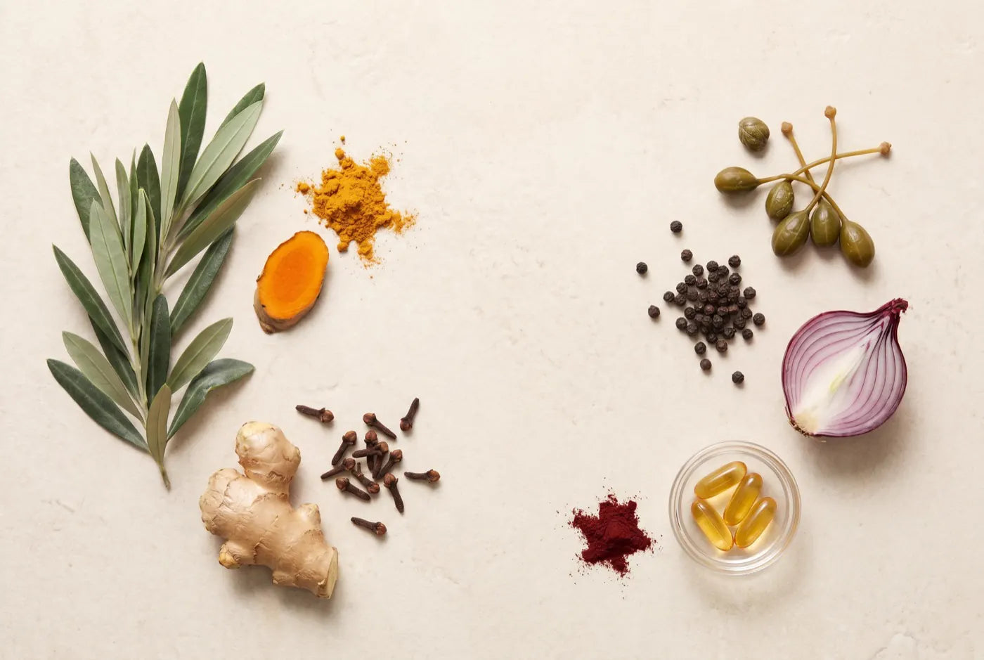 Aufsicht-Flatlay von Olivenblatt, Kurkumawurzel und -pulver, Ingwer, Nelke, Pfefferkörnern, Kapern, roter Zwiebel, astaxanthin-Pulver und goldenen Softgel-Kapseln auf warmem cremefarbenem Stein