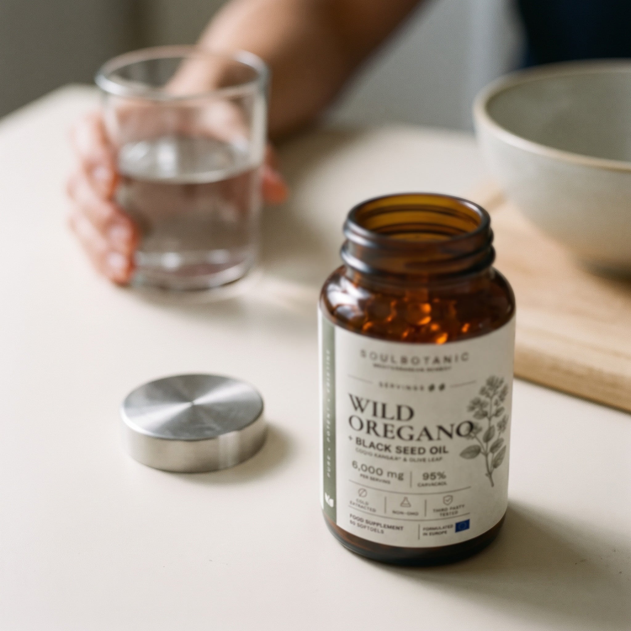 Open SoulBotanic Wild Oregano + Black Seed Oil jar on a pale kitchen table in soft daylight, its brushed steel cap resting beside it and orange softgels visible inside, with a hand lifting a glass of water blurred in the background.
